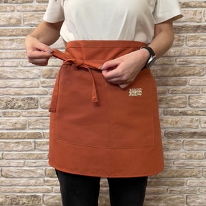 May include: A rust-colored apron with adjustable ties and a small pocket. The apron has the text "Make Make" embroidered on the front in a light yellow color.