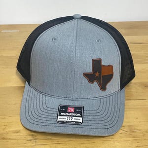 May include: A gray and black trucker hat with a brown leather Texas state patch. The patch features a white star in the center. The hat has a black mesh back and a Richardson 112 adjustable strap.