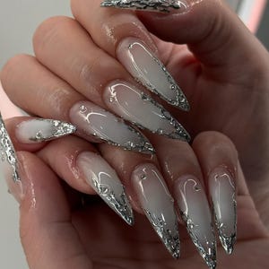 May include: Long, pointed acrylic nails with a gradient white and clear design. Each nail is adorned with silver metallic accents along the edges and small rhinestones. The nails are a fashionable accessory.