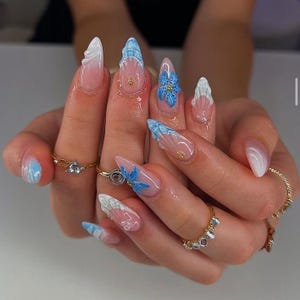 Puede incluir: Primer plano de una mano con uñas largas en forma de almendra. Las uñas presentan una base transparente con diseños blancos y azules, incluyendo patrones florales y de olas. Anillos dorados adornan los dedos, complementando el arte de uñas.