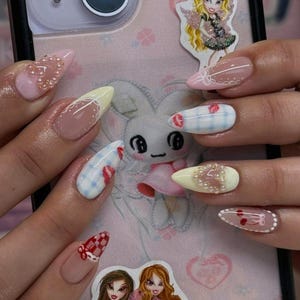 May include: A close-up of a hand with long, almond-shaped nails. The nails feature various designs, including pink, yellow, and clear bases with intricate patterns, rhinestones, and checkered accents. The background includes a phone case with a cartoon bunny and doll stickers.