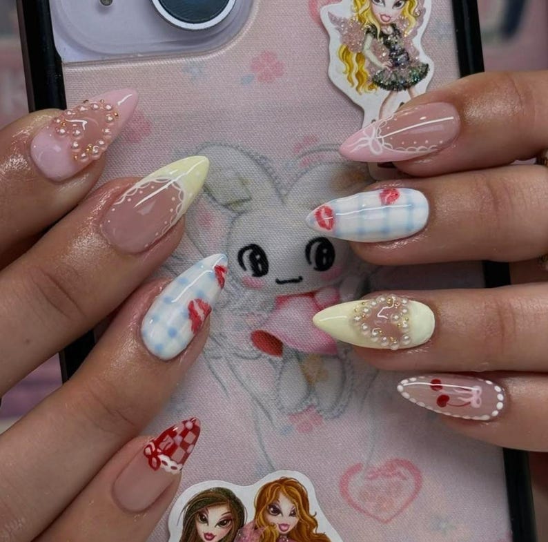 May include: A close-up of a hand with long, almond-shaped nails. The nails feature various designs, including pink, yellow, and clear bases with intricate patterns, rhinestones, and checkered accents. The background includes a phone case with a cartoon bunny and doll stickers.