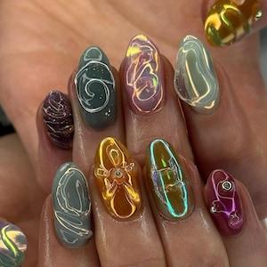 Könnte beinhalten: Nahaufnahme einer Hand mit einer Maniküre mit verschiedenen Nageldesigns. Die Nägel zeigen eine Reihe von Farben, darunter schillernd, gold und lila, mit 3D-Verzierungen und abstrakten Mustern. Die Nagelformen sind oval.