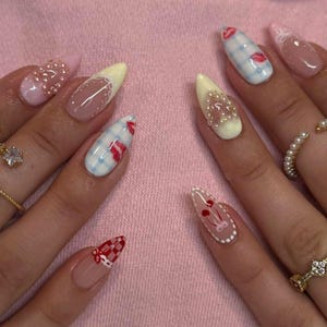 May include: A close-up of manicured nails with various designs. Some nails are painted with a light pink, yellow, and clear base, adorned with pearls, gingham patterns, and red lip and cherry designs. The nails are long and pointed.
