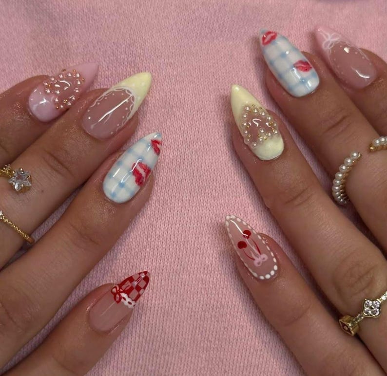 May include: A close-up of manicured nails with various designs. Some nails are painted with a light pink, yellow, and clear base, adorned with pearls, gingham patterns, and red lip and cherry designs. The nails are long and pointed.