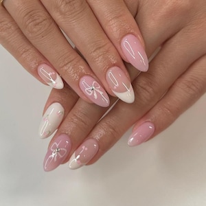 Può includere: Primo piano di unghie curate con una combinazione di rosa tenue e bianco. Alcune unghie presentano una french manicure bianca, altre un delicato design a fiocco con una piccola gemma. Le unghie ovali hanno una finitura lucida.