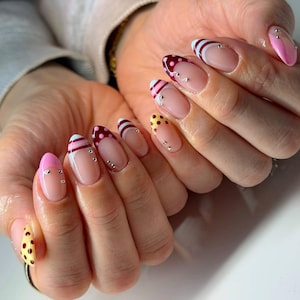 Peut inclure: Gros plan sur une main avec une manucure aux motifs variés. Certains ongles ont une base rose avec un bout blanc et des strass argentés. D'autres ongles ont une base transparente avec des pois ou des rayures rouges, jaunes et roses.