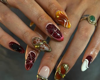 May include: A set of long, almond-shaped fingernails with various designs. Some nails feature a fruit slice design with red and pink hues, while others have abstract patterns in orange and white. Gold accents and rings are also present.