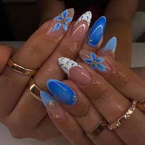 Pode incluir: Unhas em formato amendoado com base nude, detalhes florais em azul e branco, pontas francesas azuis e pequenos adornos dourados. O design inclui flores azuis pintadas à mão, pontas azuis sólidas e pontas brancas com pontos azuis, com anéis dourados.