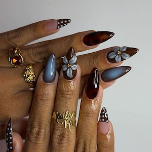 May include: Almond-shaped nails featuring a mix of brown tortoiseshell patterns, solid dark grey, and brown with light blue polka dots. Some nails are adorned with raised, light blue floral accents with gold centers. Two gold rings are worn.
