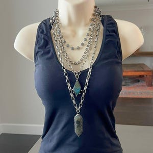 Op de afbeelding: Een marineblauwe tanktop op een mannequin, versierd met gelaagde zilveren kettingen. De kettingen hebben verschillende kettingstijlen en hangers, waaronder een druppelvormige blauwgroene steen en een rechthoekige stenen hanger.