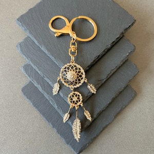 Peut inclure: Porte-clés attrape-rêves doré avec mousqueton et anneau porte-clés. Le motif attrape-rêves comporte deux cadres circulaires ornés de cristaux transparents et de breloques en forme de plumes. Le porte-clés est présenté sur une pile de dessous de plat en ardoise grise.