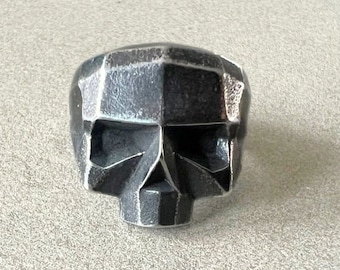 May include: A silver skull ring with a geometric design. The ring is made of metal and has a matte finish.