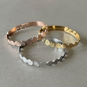 May include: Three gold bangle bracelets with a coin design. The bracelets are in rose gold, gold, and silver.