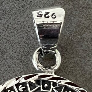 May include: Close-up of a silver pendant with a detailed geometric design. The pendant has a loop at the top and is marked with "925". The design features arrows, triangles, and other shapes, with a dark background.