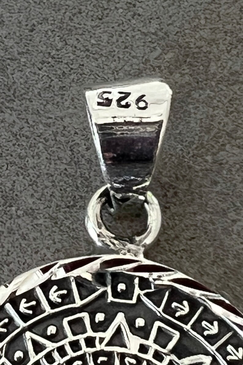 May include: Close-up of a silver pendant with a detailed geometric design. The pendant has a loop at the top and is marked with "925". The design features arrows, triangles, and other shapes, with a dark background.