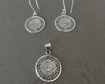 May include: A silver jewelry set featuring a pendant and matching earrings. The pendant and earrings have a circular design with an intricate pattern. The earrings have a hook closure. The jewelry is displayed on a gray surface.