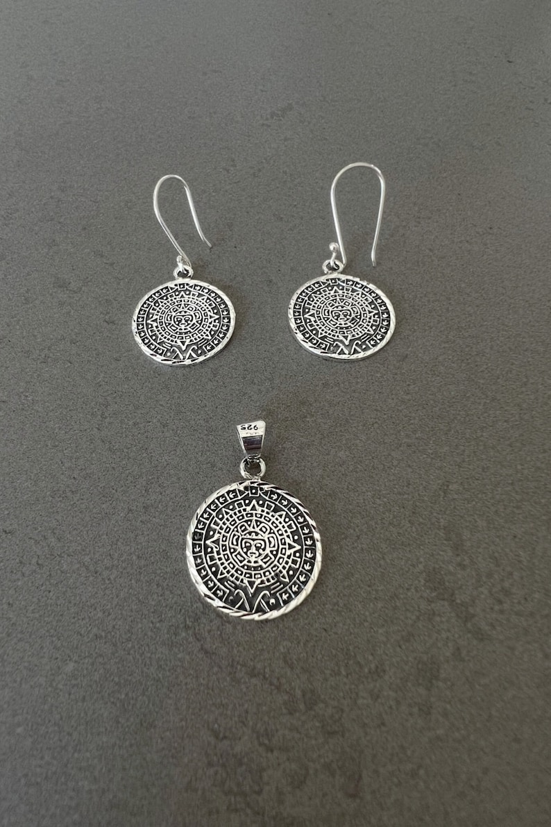 May include: A silver jewelry set featuring a pendant and matching earrings. The pendant and earrings have a circular design with an intricate pattern. The earrings have a hook closure. The jewelry is displayed on a gray surface.
