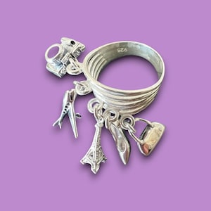 May include: A silver ring with multiple charms hanging from it. The charms include a small airplane, a miniature Eiffel Tower, a shoe, a handbag, and a camera. The ring is marked with the number "925".