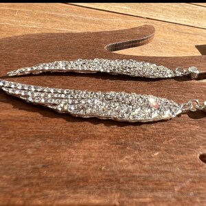 May include: A pair of silver earrings shaped like angel wings, each wing is covered in clear rhinestones.