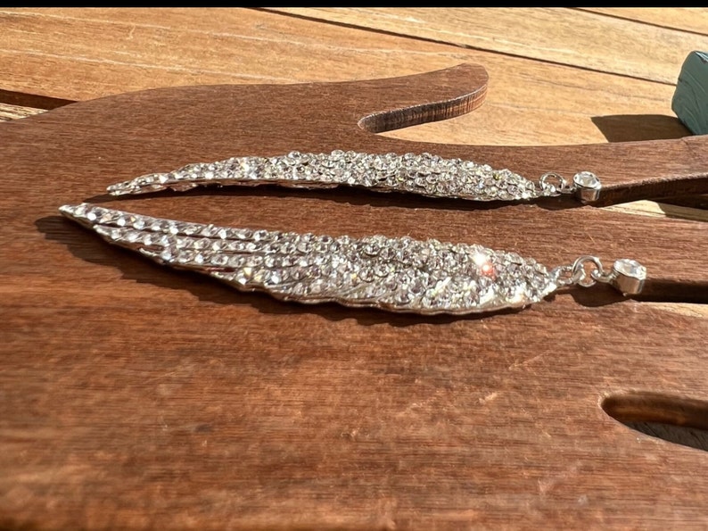 May include: A pair of silver earrings shaped like angel wings, each wing is covered in clear rhinestones.
