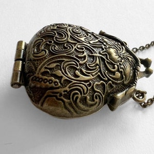 Puede incluir: Un collar con un medall&oacute;n de bronce, con una tapa con bisagras y un dise&ntilde;o floral detallado. El medall&oacute;n es de forma ovalada y tiene una cadena.