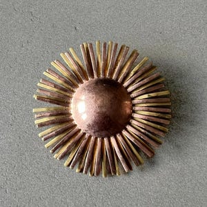 May include: A round, sunburst-style brooch with a copper-colored center and radiating bars in alternating copper and gold tones. The brooch is set against a gray background.