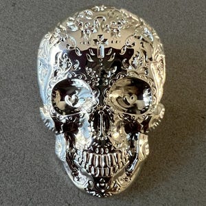May include: A silver-colored, decorative skull with intricate floral and cross designs. The skull features heart-shaped eye sockets and detailed carvings, reflecting light. This Day of the Dead-themed item is likely a decorative piece.