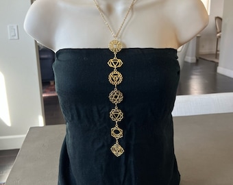May include: A gold chain necklace with seven chakra symbols hanging from it. The symbols are in a vertical line and are spaced evenly apart. The necklace is on a black mannequin.