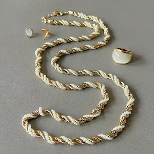 May include: A gold-tone rope necklace with white detailing, a matching ring, and a pair of earrings. The jewellery is displayed on a grey surface. The ring has a white face with a gold band.
