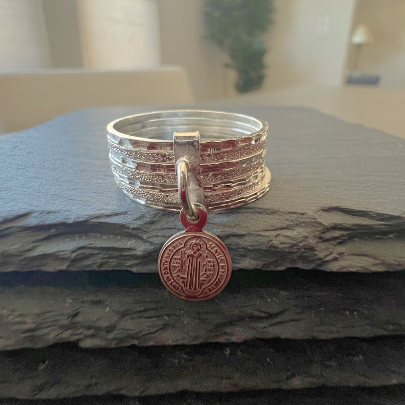 May include: A silver ring with a charm of Saint Anthony of Padua. The ring is made of multiple bands with a textured finish.