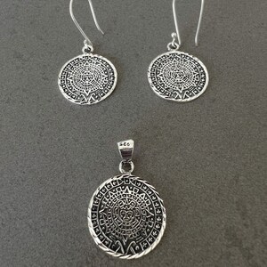 May include: A silver pendant necklace and matching earrings set. The jewelry features a circular design with an intricate pattern. The earrings have a hook closure. The pendant is marked with "925".