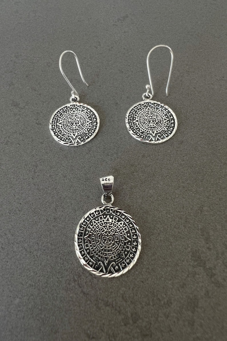May include: A silver pendant necklace and matching earrings set. The jewelry features a circular design with an intricate pattern. The earrings have a hook closure. The pendant is marked with "925".
