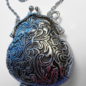 Puede incluir: Un collar de plata con un colgante en forma de bolsa adornado con detalles. El colgante tiene un dise&ntilde;o floral y una abertura con bisagra.