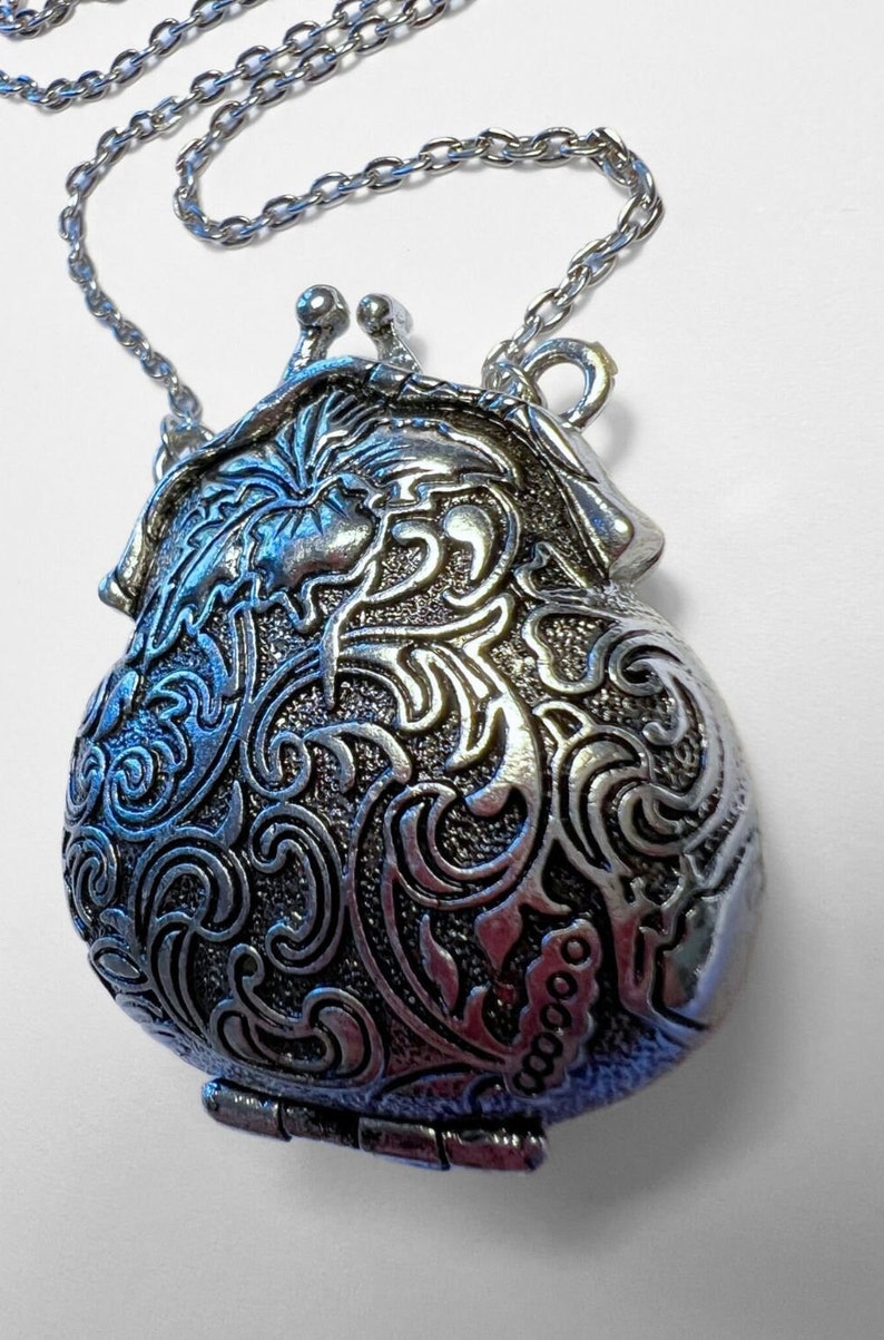Puede incluir: Un collar de plata con un colgante en forma de bolsa adornado con detalles. El colgante tiene un dise&ntilde;o floral y una abertura con bisagra.