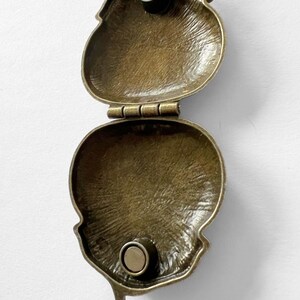 Puede incluir: Un collar con un colgante de medall&oacute;n de color bronce, con una tapa con bisagra. El medall&oacute;n est&aacute; abierto, revelando un interior liso y vac&iacute;o. El medall&oacute;n est&aacute; unido a una cadena.