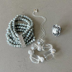 May include: A selection of jewellery including a multi-strand beaded bracelet with a rhinestone clasp, a silver necklace with clear crystal beads, and a silver ring with a large, clear stone. The bracelet and necklace are silver and clear.