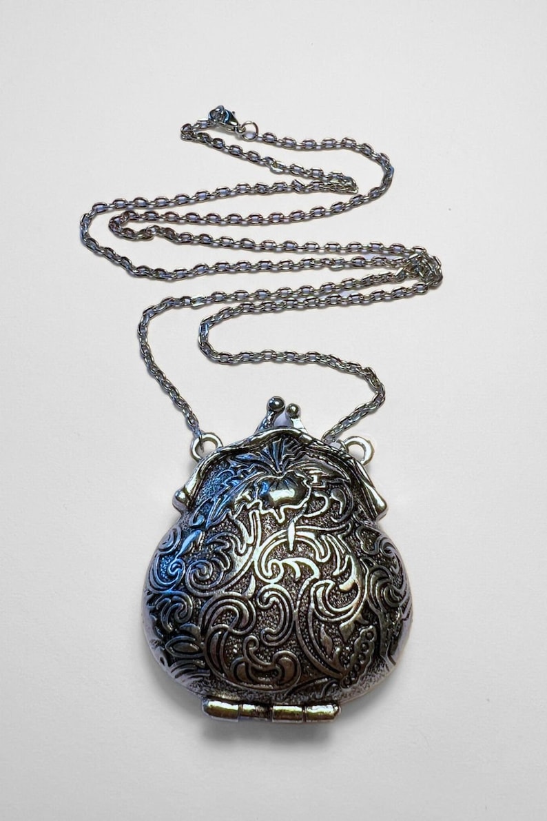 Puede incluir: Un collar de cadena de plata con un colgante de medall&oacute;n de plata. El medall&oacute;n tiene forma de bolso y presenta un dise&ntilde;o floral ornamentado.