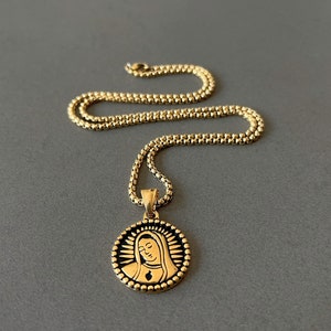 May include: Gold-colored necklace with a round pendant featuring a religious figure. The pendant is encircled by a beaded border and has a detailed image. The chain is a textured, box-style design. The necklace is displayed on a gray surface.