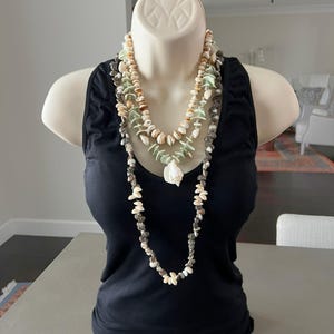 May include: A black sleeveless tank top worn by a mannequin, layered with two distinct necklaces. The shorter necklace features small tan and light green shells, while the longer necklace consists of small grey and white shells in a long strand.