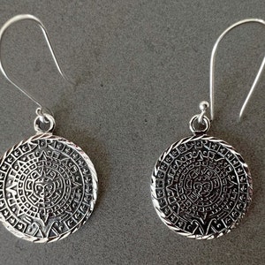 May include: A pair of silver-colored dangle earrings. Each earring features a circular pendant with an intricate, detailed design reminiscent of an ancient calendar or sun symbol. The earrings have a hook closure.