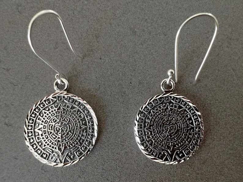 May include: A pair of silver-colored dangle earrings. Each earring features a circular pendant with an intricate, detailed design reminiscent of an ancient calendar or sun symbol. The earrings have a hook closure.