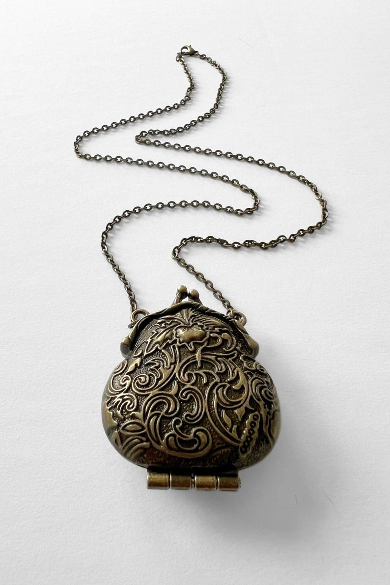 Puede incluir: Un collar dorado con un colgante de medall&oacute;n detallado, adornado y con bisagras. El medall&oacute;n tiene forma de bolsa y presenta un dise&ntilde;o floral.
