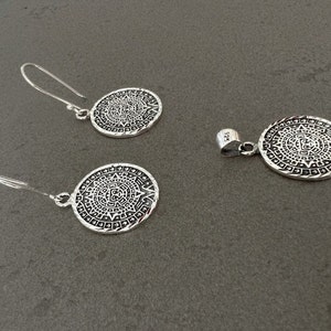 May include: A silver jewelry set featuring a pendant and a pair of earrings. Each piece has a circular design with an intricate pattern. The earrings have a hook closure. The pendant has a loop for a chain. The set is made of a shiny, metallic material.