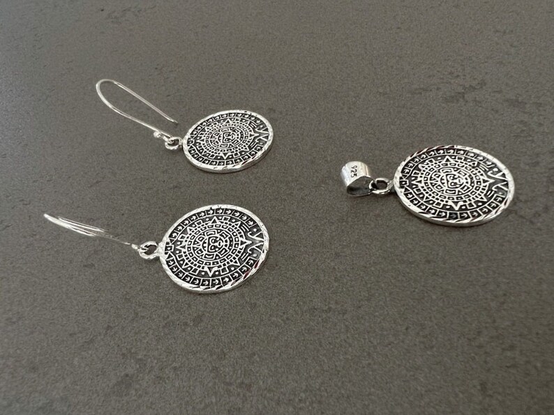 May include: A silver jewelry set featuring a pendant and a pair of earrings. Each piece has a circular design with an intricate pattern. The earrings have a hook closure. The pendant has a loop for a chain. The set is made of a shiny, metallic material.