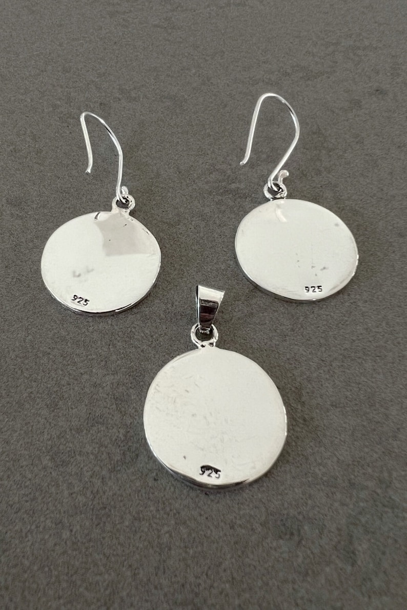 May include: A silver jewelry set featuring a pendant and a pair of earrings. Each piece has a circular design with a smooth, reflective surface. The earrings have hook closures, and all pieces are marked with "925".