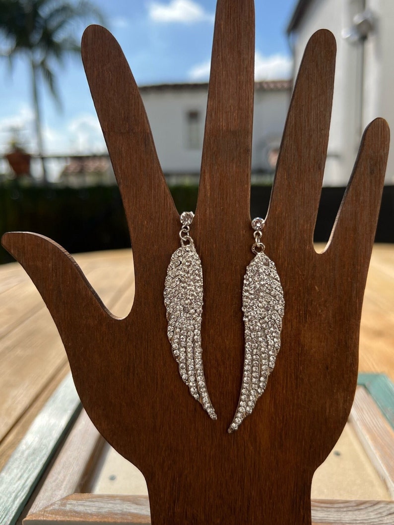 May include: A pair of silver dangle earrings with clear crystal wings. The earrings are on a wooden display stand.