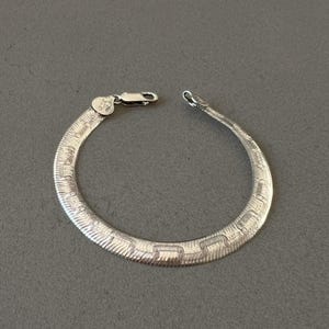 May include: A silver herringbone bracelet with a lobster clasp. The bracelet is made of a series of interlocking, textured links. The clasp has a small, decorative element. The bracelet is displayed on a gray surface.