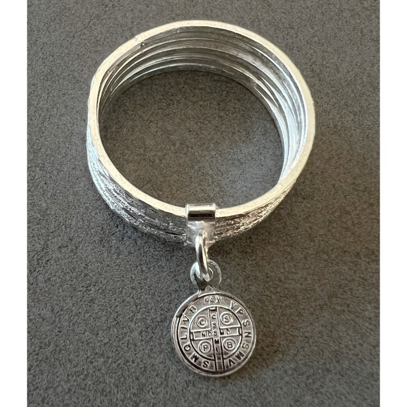 May include: Silver ring with a charm of a Saint Benedict medal. The ring is made of five bands.