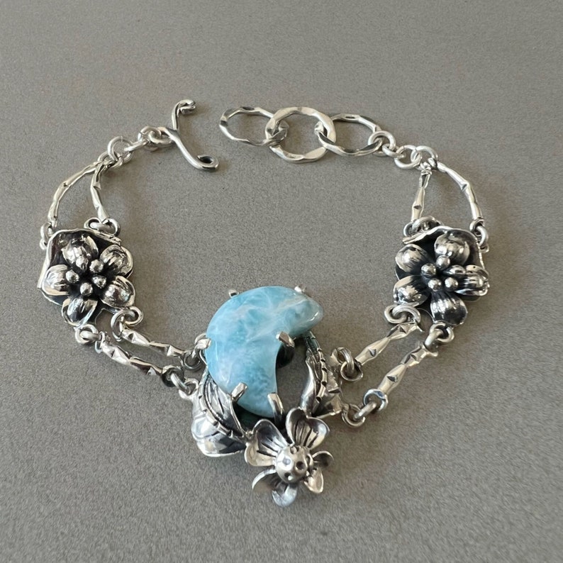 以下が含まれることがあります： 青い宝石と花柄のアクセントが付いたシルバーブレスレット。ブレスレットは繊細なチェーンと留め金で留められています。